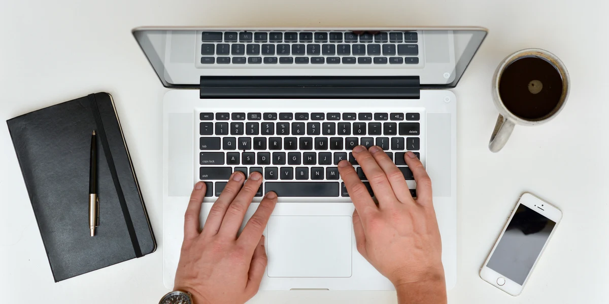 Overhead view of two hands resting on a laptop keyboard on a white desk, next to a notebook, a smartphone and a cup of coffee