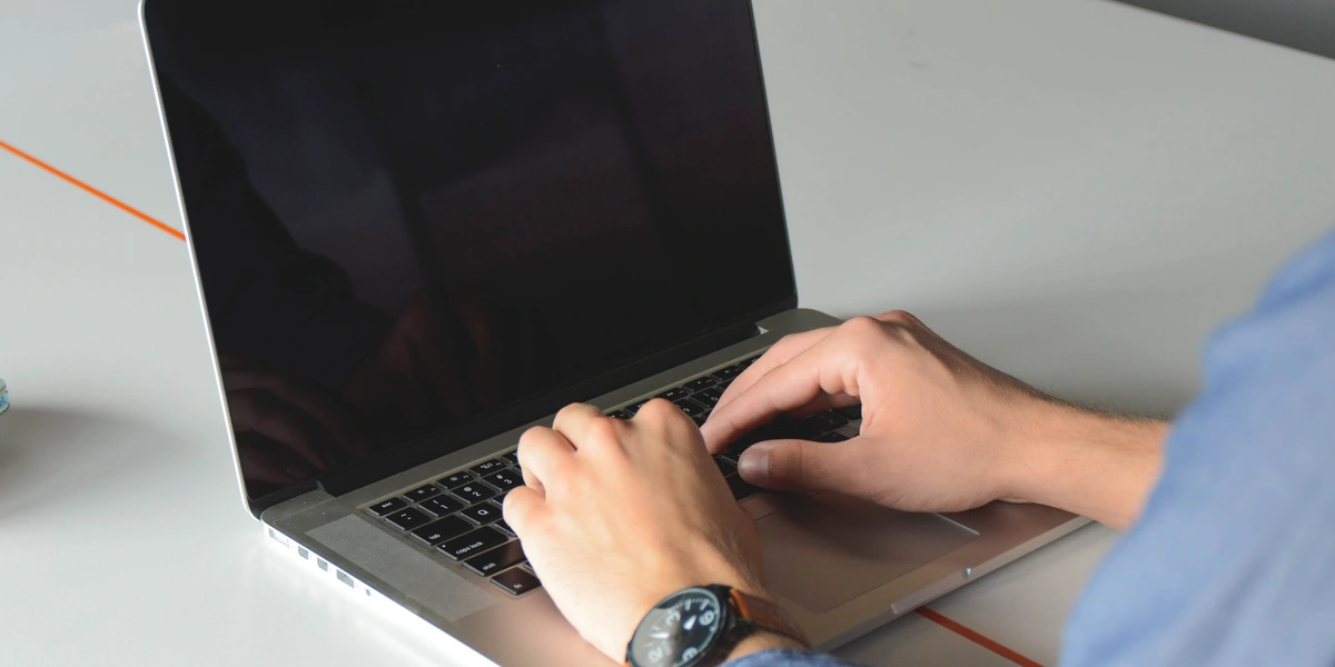 A person in a light blue denim shirt typing on an open laptop at a bright white desk